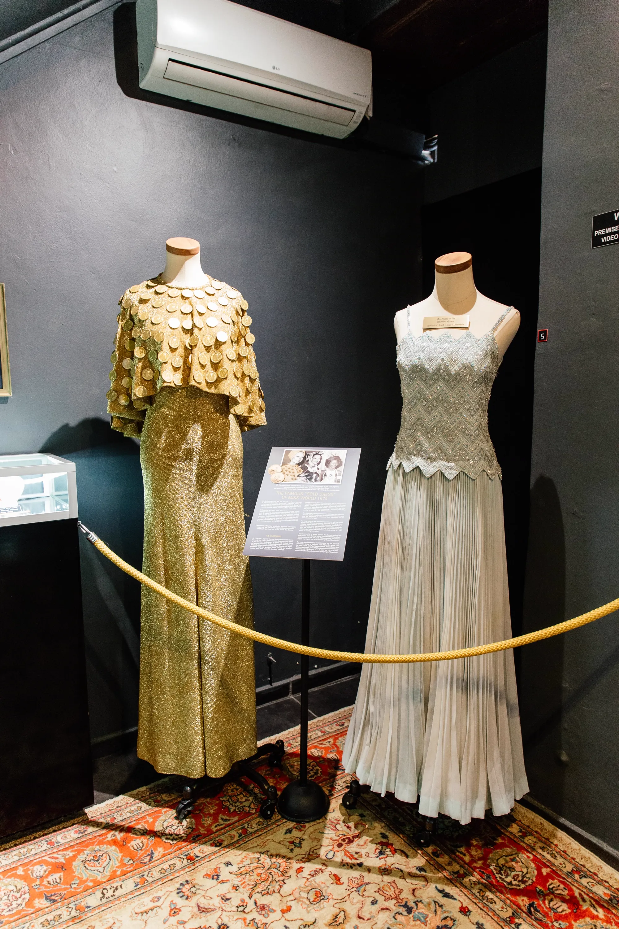 The gallery's gold-rope-cordoned plinth room in the museum: the Miss World 1974 Krugerrand gown on the left mannequin, Anneline Kriel's silver beaded evening gown on the right, both on a Persian rug under warm gallery lighting