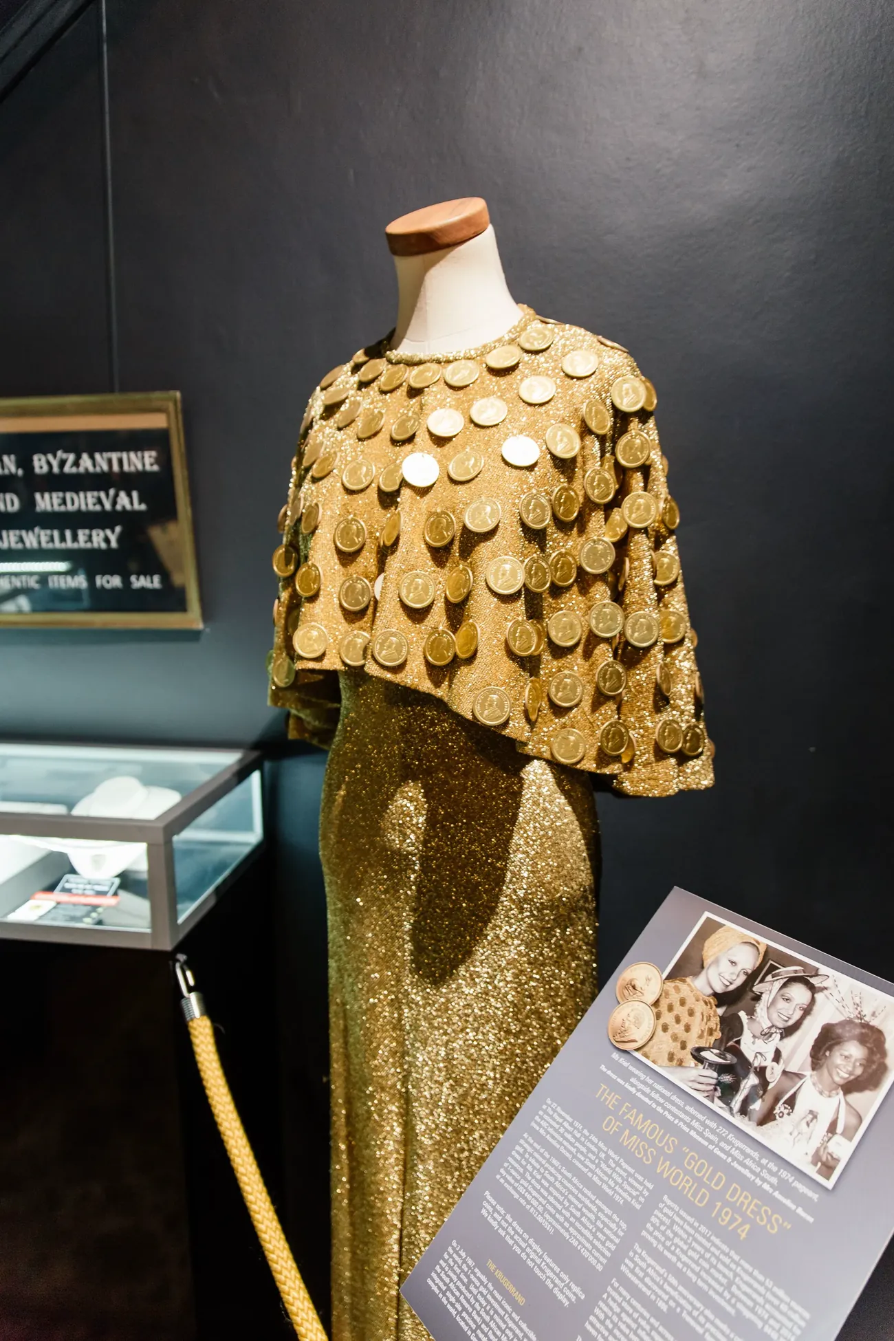 The Miss World 1974 Krugerrand gown on display in the museum, adorned with 272 replica Krugerrand coins, with the archival photograph of Anneline Kriel and the story plaque beside it