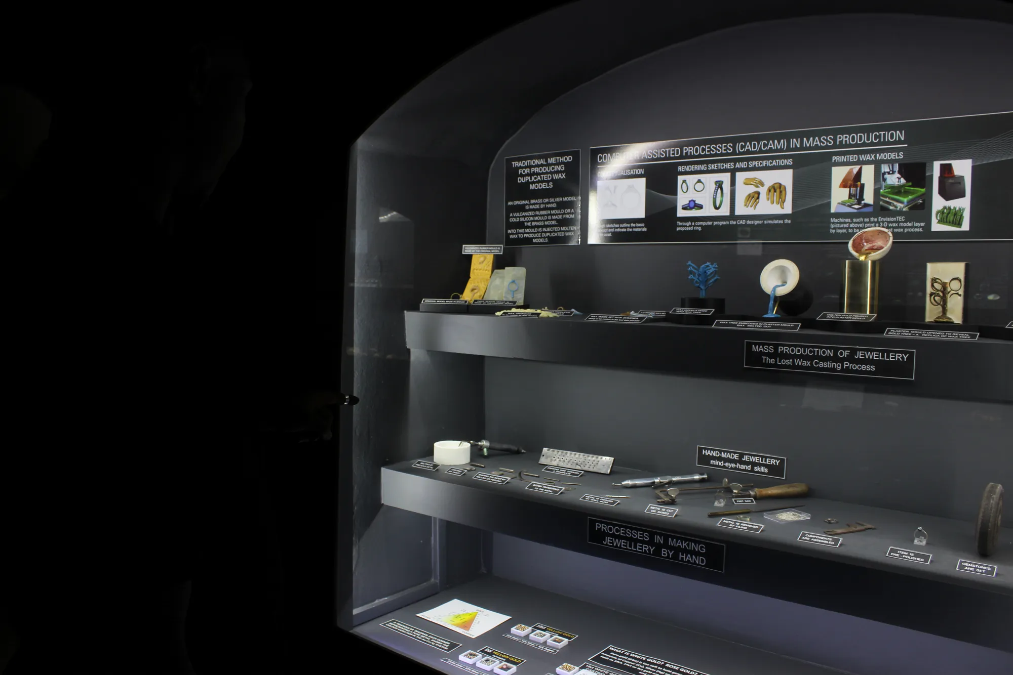 The Making Jewellery cabinet in the museum: the lost-wax casting process from CAD render through 3-D printed wax model, wax tree, plaster mould, molten gold pour and final polished ring, shown alongside the hand-made jewellery shelf with goldsmith's drawplate, files and tools