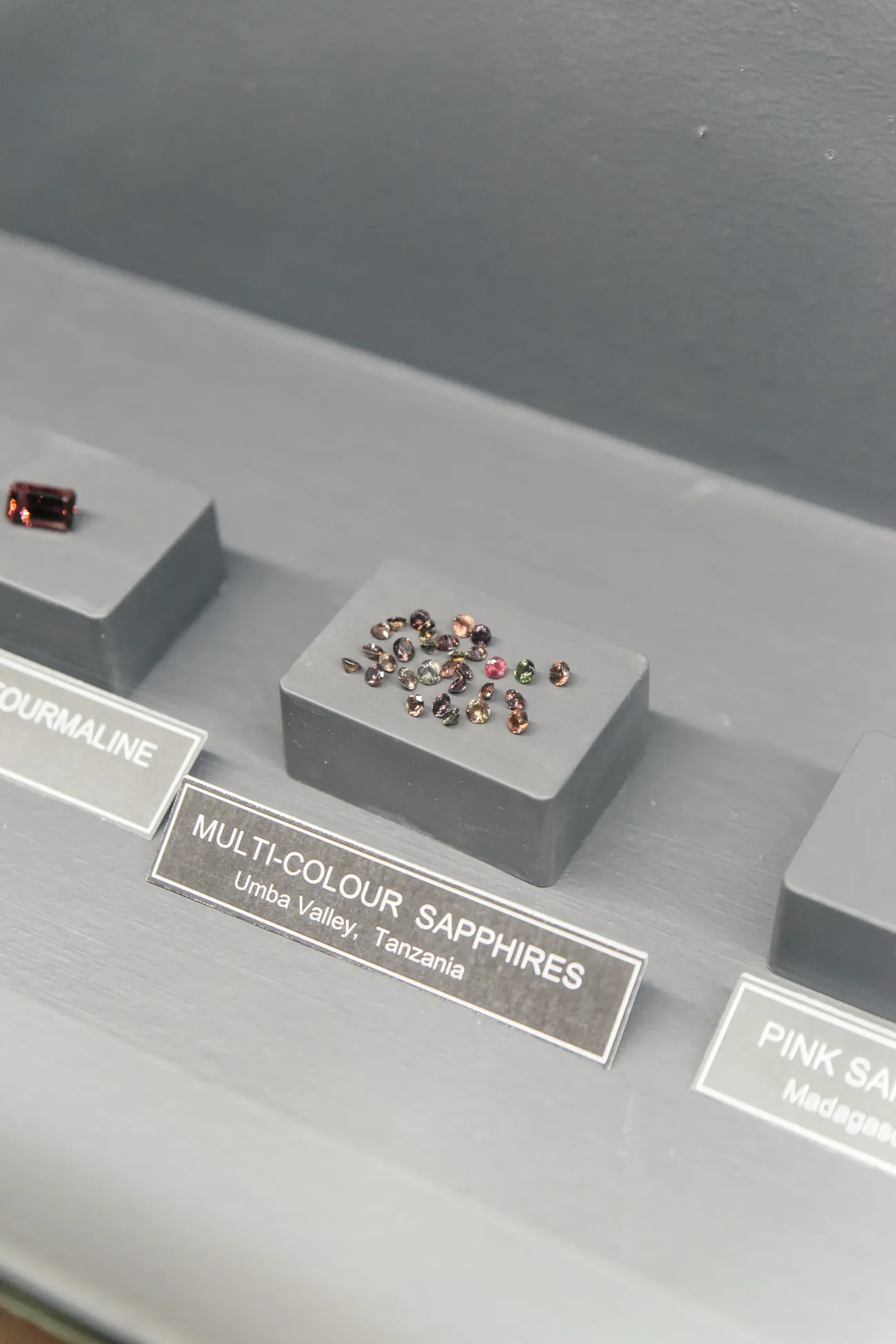 A parcel of multi-colour sapphires from the Umba Valley, Tanzania, on a black plinth in the museum, beside an emerald-cut tourmaline