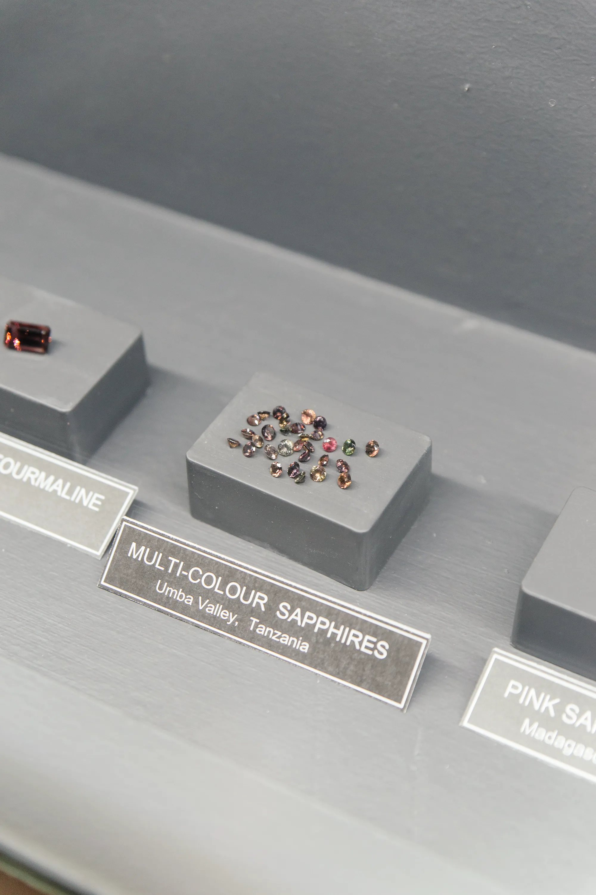 A parcel of multi-colour sapphires from the Umba Valley, Tanzania, on a black plinth in the museum, beside an emerald-cut tourmaline