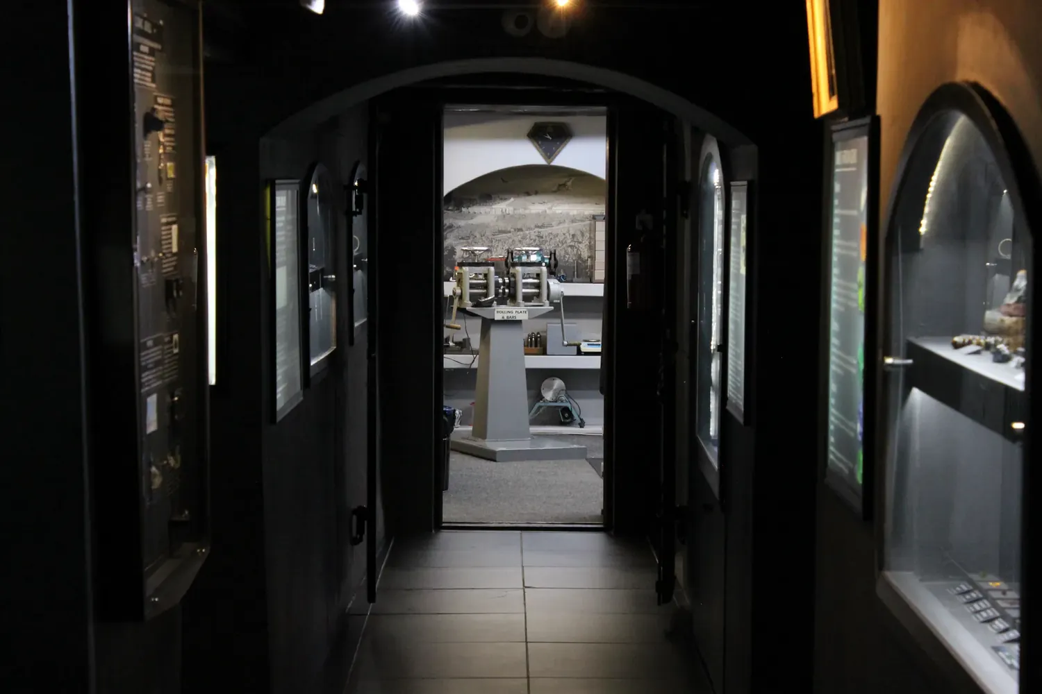 A corridor of lit display cases in the Museum of Gems & Jewellery leading through an arched doorway to a room of goldsmith’s equipment, with rolling mills for plate and bars at its centre